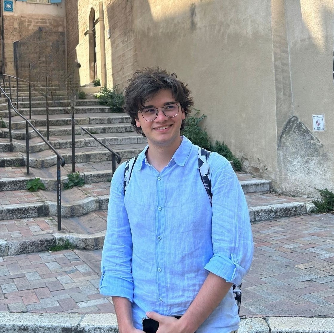 Riccardo Ali smiling outdoors, wearing glasses and a blue shirt